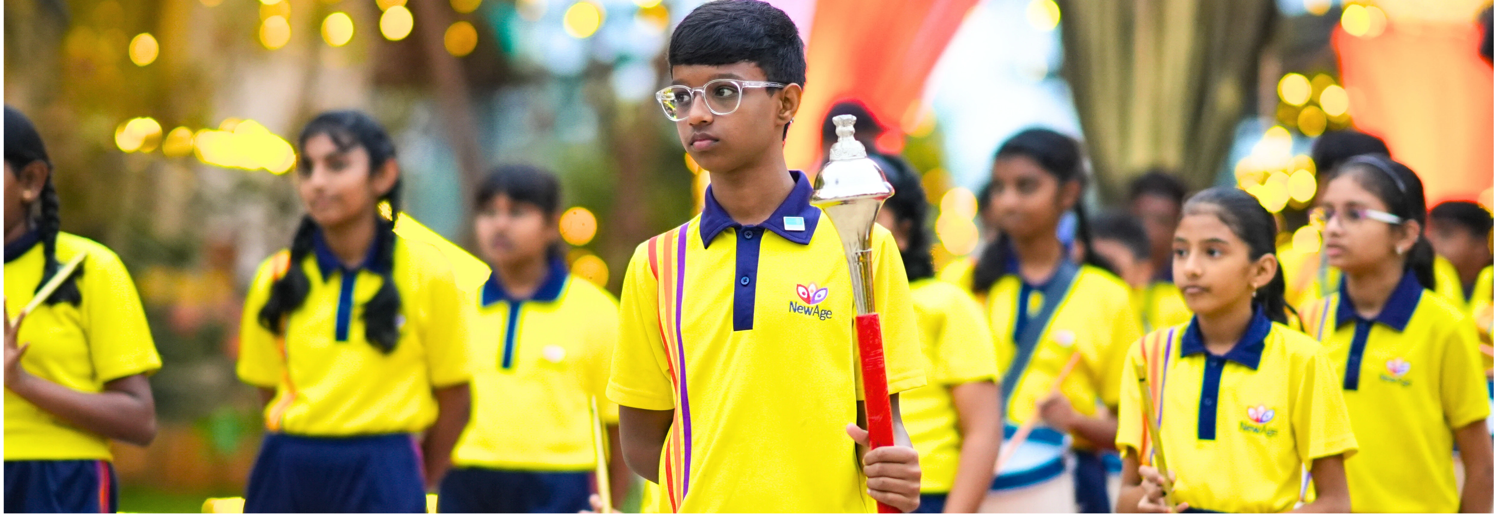 Students of NewAge World School participating in a ceremonial march, dressed in school uniforms, with a student leading the procession holding a torch.